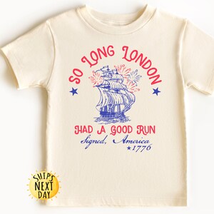 May include: Cream-colored t-shirt featuring a blue and red graphic of a tall ship with fireworks and the text "SO LONG LONDON HAD A GOOD RUN Signed, America *1776". The shirt is made of soft material.