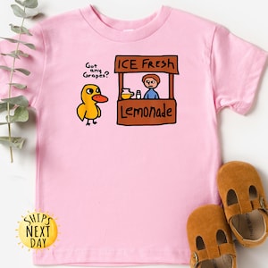 May include: Pink short-sleeved t-shirt featuring a cartoon duck asking "Got any Grapes?" next to a lemonade stand that says "ICE FRESH Lemonade." Brown leather baby shoes are at the bottom of the image.