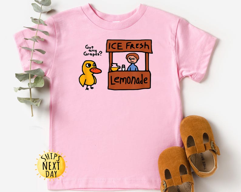 May include: Pink short-sleeved t-shirt featuring a cartoon duck asking "Got any Grapes?" next to a lemonade stand that says "ICE FRESH Lemonade." Brown leather baby shoes are at the bottom of the image.