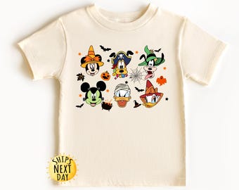 May include: A cream-coloured t-shirt with a Halloween-themed design featuring Mickey Mouse, Donald Duck, Goofy, and Pluto dressed in costumes. The characters are wearing hats and have pumpkins and bats around them.