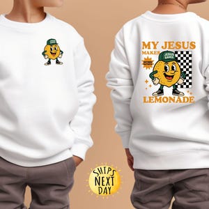 May include: White sweatshirt featuring a cartoon lemon character wearing a green cap and sneakers. The front has a small lemon graphic, while the back displays the text "MY JESUS MAKES LEMONADE" with the lemon character and a checkered background.