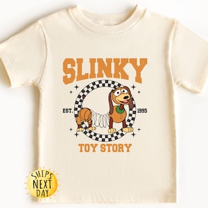 May include: A cream-colored t-shirt with a graphic of Slinky Dog from Toy Story. The graphic features Slinky Dog in a checkered circle with the text "SLINKY" above and "EST. 1995" below. The text "TOY STORY" is below the circle.