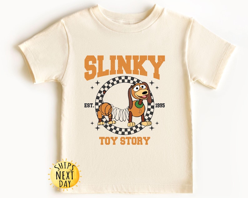 May include: A cream-colored t-shirt with a graphic of Slinky Dog from Toy Story. The graphic features Slinky Dog in a checkered circle with the text "SLINKY" above and "EST. 1995" below. The text "TOY STORY" is below the circle.