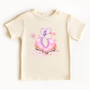 May include: Cream-colored t-shirt featuring a graphic of pink roller skates with gold accents and a purple heart-shaped design. The skates are adorned with floral and star details. A whimsical design for children's apparel.