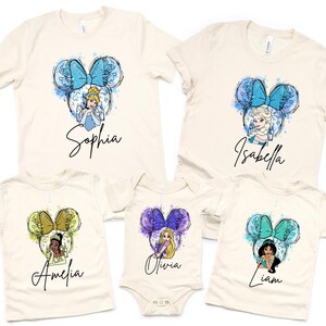 May include: A collection of five cream-colored t-shirts and one bodysuit featuring watercolor-style graphics of Disney characters inside mouse ear silhouettes. Each garment displays a unique character, a colorful bow, and a personalized name in script.