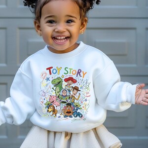 May include: A white long-sleeve sweatshirt featuring a colorful graphic print of Toy Story characters including Woody, Buzz Lightyear, Rex, and Jessie. The design includes the text Toy Story and scribbled crayon-style drawings in various colors.