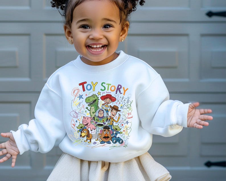 May include: A white long-sleeve sweatshirt featuring a colorful graphic print of Toy Story characters including Woody, Buzz Lightyear, Rex, and Jessie. The design includes the text Toy Story and scribbled crayon-style drawings in various colors.