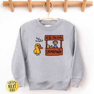 May include: A light gray sweatshirt featuring a cartoon duck asking "Got any Grapes?" next to a lemonade stand with the text "ICE FRESH Lemonade." The design is in shades of yellow, orange, brown, and blue.