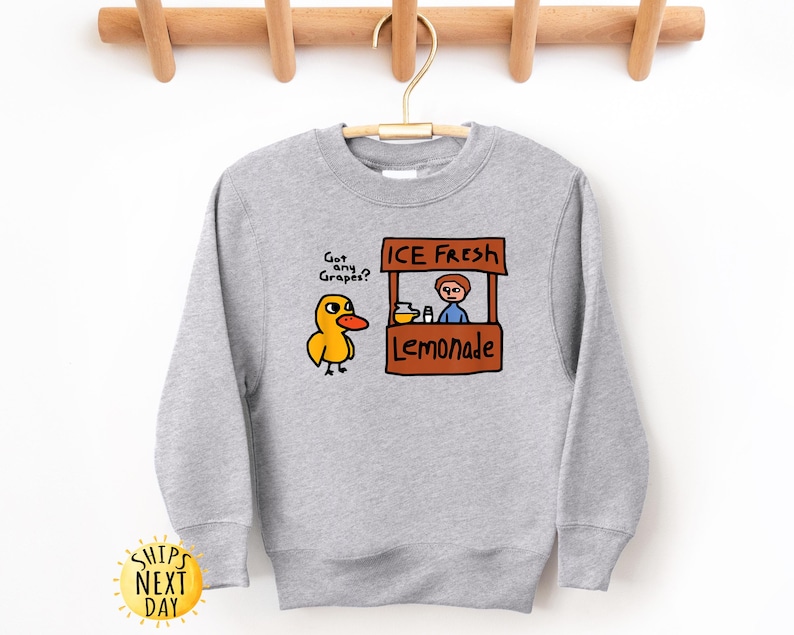 May include: A light gray sweatshirt featuring a cartoon duck asking "Got any Grapes?" next to a lemonade stand with the text "ICE FRESH Lemonade." The design is in shades of yellow, orange, brown, and blue.
