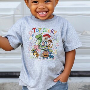 May include: A light gray short-sleeve t-shirt featuring a colorful graphic print of Toy Story characters including Woody, Buzz Lightyear, Rex, and Mr. Potato Head, with the text Toy Story and scribbled star and swirl designs in various colors.