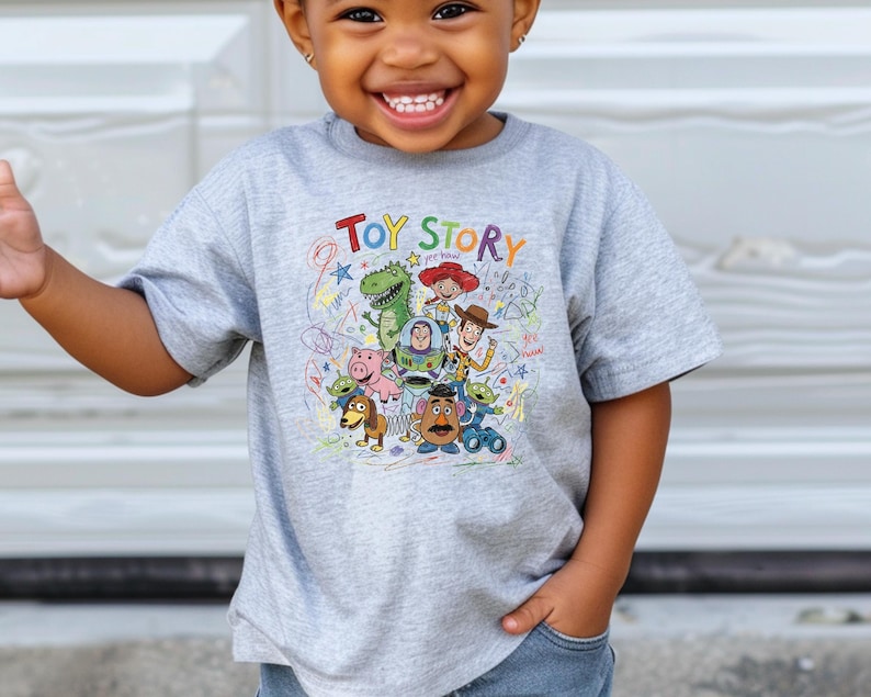 May include: A light gray short-sleeve t-shirt featuring a colorful graphic print of Toy Story characters including Woody, Buzz Lightyear, Rex, and Mr. Potato Head, with the text Toy Story and scribbled star and swirl designs in various colors.