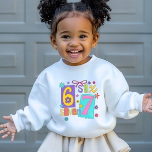 May include: A white sweatshirt with the numbers "6" and "7" in colorful boxes, along with the words "six" and "seven". The sweatshirt has a floral and star design. The child is smiling and wearing a skirt.