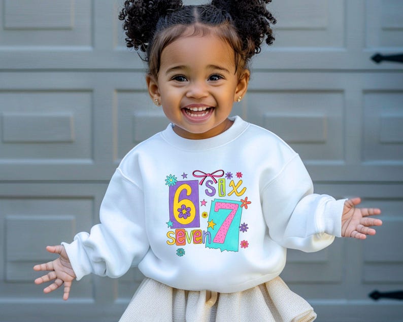 May include: A white sweatshirt with the numbers "6" and "7" in colorful boxes, along with the words "six" and "seven". The sweatshirt has a floral and star design. The child is smiling and wearing a skirt.