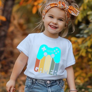 May include: White t-shirt featuring a teal video game controller graphic with the word "GAMER" in a colorful retro design. The shirt is worn by a young child.