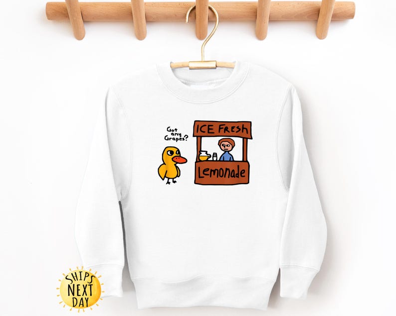 May include: White sweatshirt with a cartoon duck asking "Got any Grapes?" next to a lemonade stand that says "ICE Fresh Lemonade." The sweatshirt has long sleeves and a crew neck. A yellow sun graphic with the words "SHIPS NEXT DAY" is on the bottom left.