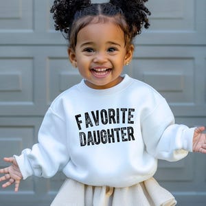 May include: A white sweatshirt with the words "FAVORITE DAUGHTER" in black lettering. The sweatshirt is worn by a young child with curly hair. The child is smiling and wearing a beige skirt.