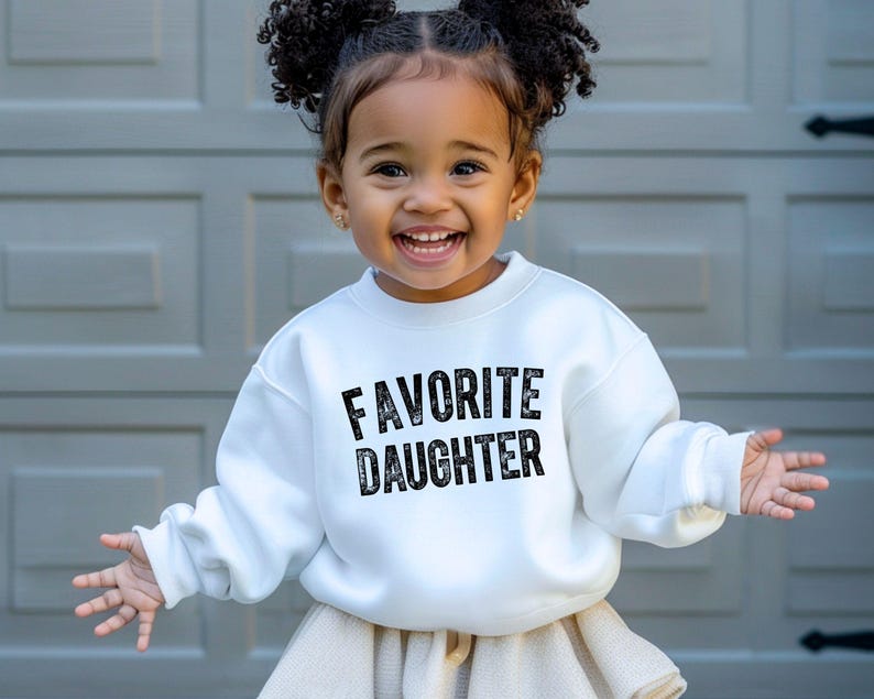 May include: A white sweatshirt with the words "FAVORITE DAUGHTER" in black lettering. The sweatshirt is worn by a young child with curly hair. The child is smiling and wearing a beige skirt.