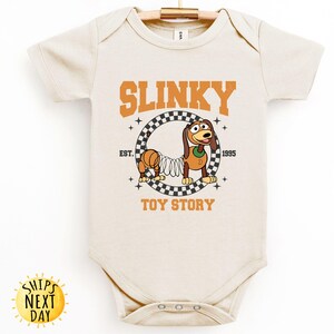 May include: A cream-colored baby onesie with a graphic of Slinky Dog from Toy Story. The graphic features Slinky Dog in a checkered circle with the text "Slinky" above and "Toy Story" below. The text "Est. 1995" is to the right of the circle.