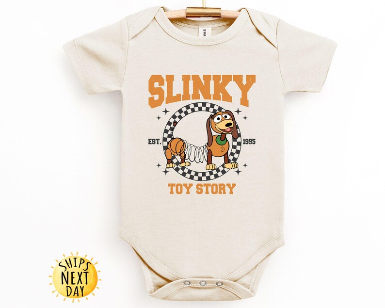 May include: A cream-colored baby onesie with a graphic of Slinky Dog from Toy Story. The graphic features Slinky Dog in a checkered circle with the text "Slinky" above and "Toy Story" below. The text "Est. 1995" is to the right of the circle.