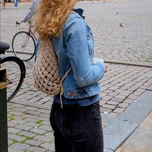 Puede incluir: Una persona con una chaqueta vaquera azul claro, vaqueros negros y una mochila de macramé beige. La persona está de pie en una calle adoquinada, con una bicicleta y edificios al fondo.