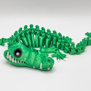 May include: A green 3D printed alligator skeleton toy with white teeth and eyes. The alligator is posed in a crawling position with its tail curled up.