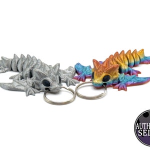Peut inclure: Deux porte-clés en forme de lézard imprimés en 3D, l'un argenté et l'autre avec une peinture arc-en-ciel irisée. Les deux lézards ont des dos hérissés et sont attachés à des anneaux de clés argentés.