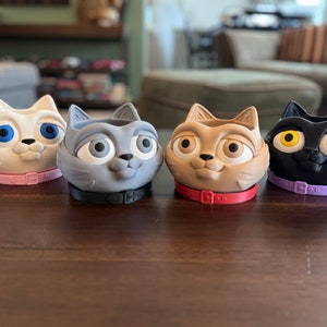 Puede incluir: Cuatro macetas de cerámica con forma de cabeza de gato con collares de colores. Las macetas son blancas, grises, marrones y negras. Cada maceta tiene ojos grandes y expresivos.