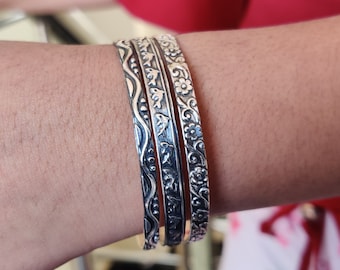 May include: Four silver bangles with intricate designs are stacked on a wrist. The bangles feature floral, wave, and leaf patterns. The silver metal reflects light, highlighting the detailed carvings. The background is blurred, focusing attention on the jewelry.