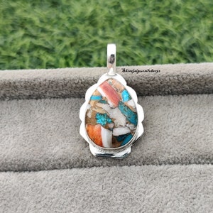 Pu&ograve; includere: Un ciondolo a forma di lacrima con una montatura in argento e un'incisione di pietra colorata. La pietra &egrave; un mix di turchese, corallo e bianco, con un motivo maculato. Il ciondolo &egrave; su uno sfondo grigio.
