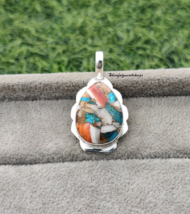 Pu&ograve; includere: Un ciondolo a forma di lacrima con una montatura in argento e un'incisione di pietra colorata. La pietra &egrave; un mix di turchese, corallo e bianco, con un motivo maculato. Il ciondolo &egrave; su uno sfondo grigio.