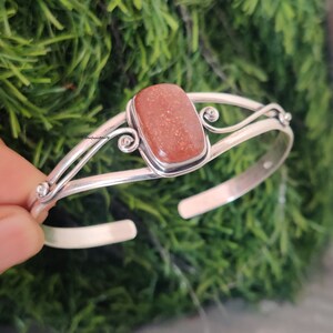 May include: A silver bangle bracelet with a rectangular, orange sunstone gemstone set in the center. The bracelet has a delicate, ornate design with swirls and curves.