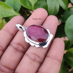 May include: A silver pendant with a large, oval-shaped ruby gemstone. The ruby is a deep red color and has a faceted cut. The pendant is set in a silver frame with a decorative border.