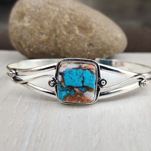 Könnte beinhalten: Ein silbernes Armband mit einem quadratischen Türkis- und Orangenstein, der in der Mitte eingesetzt ist. Das Armband hat ein Doppelband-Design mit einem Wirbeldetail auf jeder Seite.
