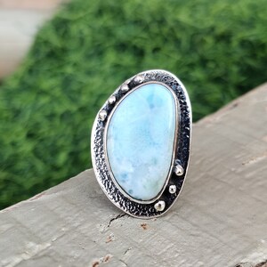 May include: A silver ring with a light blue, oval-shaped gemstone set in a textured silver bezel. The ring has a hammered finish and small silver beads around the edge of the bezel.