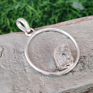 May include: A silver pendant with a clear, faceted crystal set in a circle. The pendant is on a wooden surface.