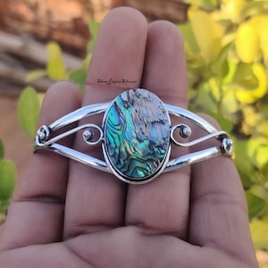 Puede incluir: Un brazalete de plata con una gran incrustación ovalada de concha de abulón. La concha tiene colores iridiscentes verdes, azules y marrones. El brazalete tiene un diseño delicado y curvo.