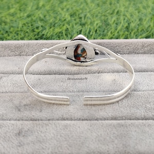 May include: Silver bangle bracelet with a colorful stone inlay. The stone is a teardrop shape and has a variety of colors, including blue, green, red, and white.