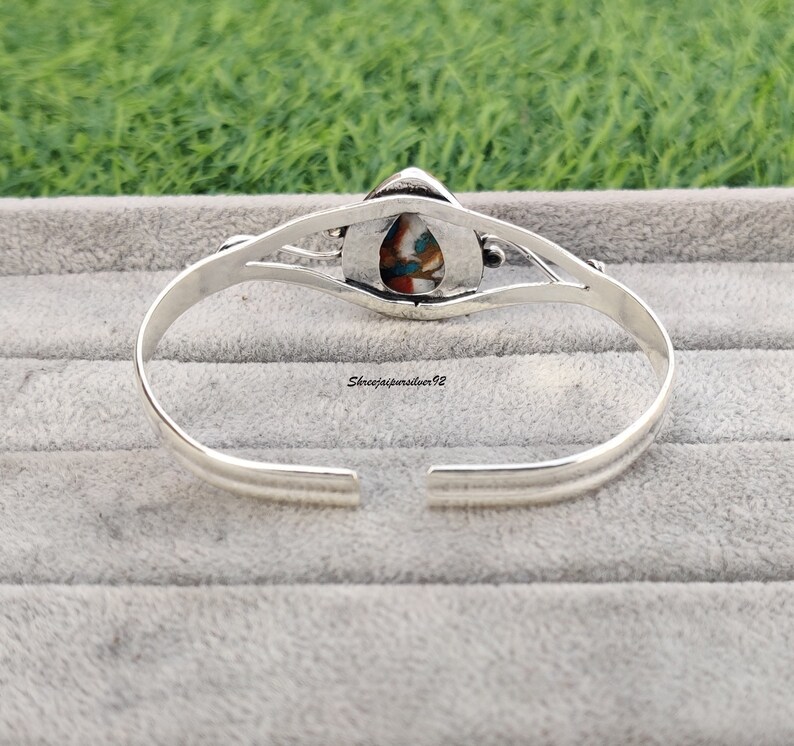 May include: Silver bangle bracelet with a colorful stone inlay. The stone is a teardrop shape and has a variety of colors, including blue, green, red, and white.