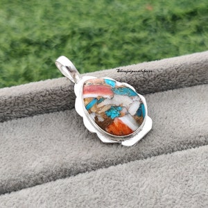 Pu&ograve; includere: Un ciondolo d'argento con una pietra ovale colorata incastonata in una cornice d'argento. La pietra &egrave; un mix di turchese, bianco, arancione e marrone.