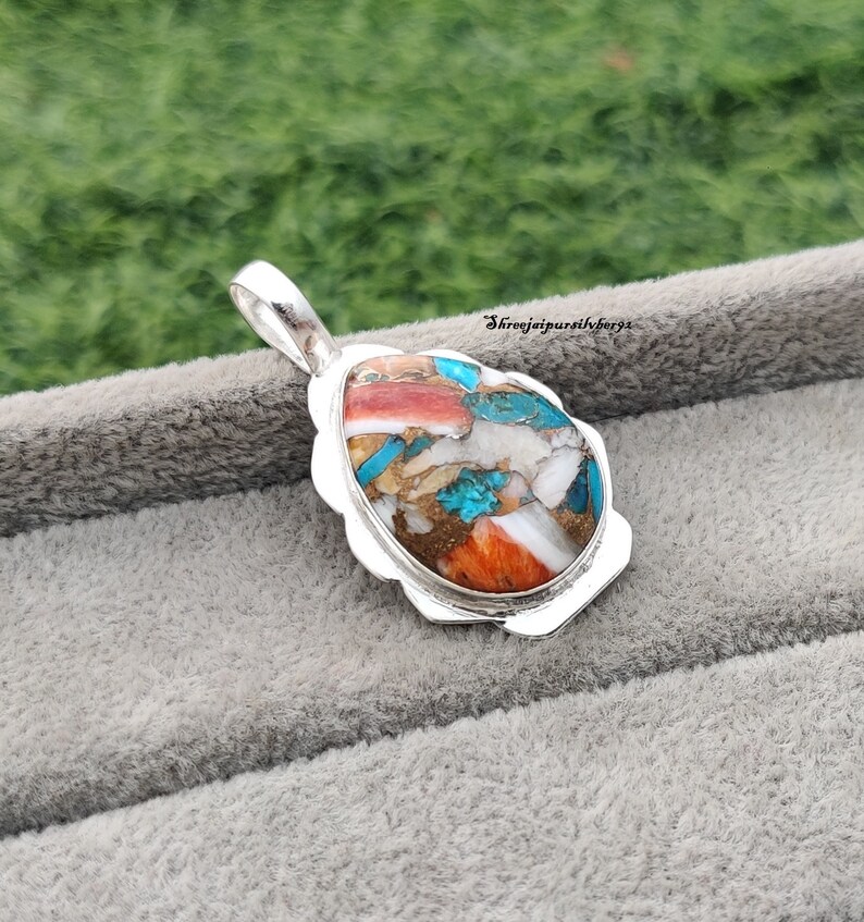 Pu&ograve; includere: Un ciondolo d'argento con una pietra ovale colorata incastonata in una cornice d'argento. La pietra &egrave; un mix di turchese, bianco, arancione e marrone.