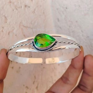 May include: A silver bangle bracelet featuring a green gemstone at its centre. The bracelet has a twisted wire design and a smooth finish.