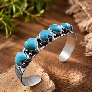 May include: A silver cuff bracelet featuring four polished, heart-shaped turquoise stones set in a row. The metal band displays engraved sunburst patterns on the sides, with a smooth, open-ended design for a comfortable fit on the wrist.