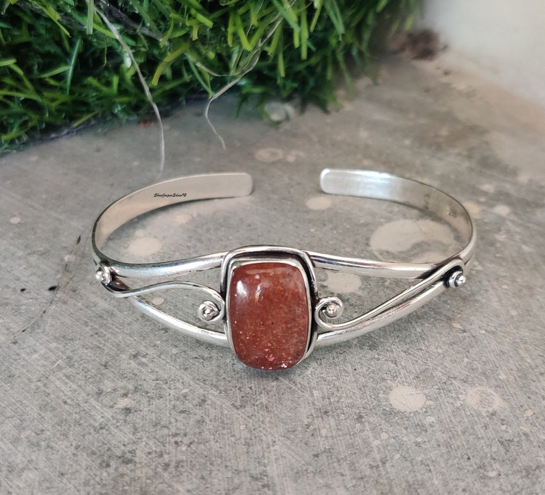 May include: Silver cuff bracelet with a rectangular, reddish-brown gemstone. The bracelet has a delicate, twisted design and is stamped "Sterling Silver".