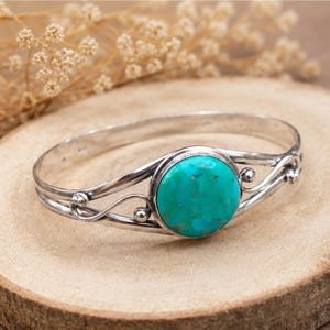 May include: A silver cuff bracelet featuring a central round turquoise stone with natural brown matrix patterns. The band is crafted from polished silver wire that curves into decorative swirls and small spheres on both sides of the stone.