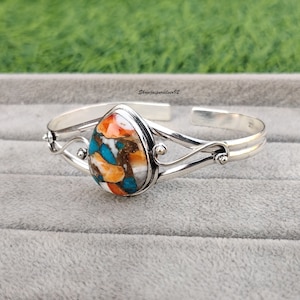 May include: A silver cuff bracelet with a teardrop-shaped turquoise stone set in the center. The stone is a mix of orange, blue, and white colors. The bracelet has a delicate, swirling design.