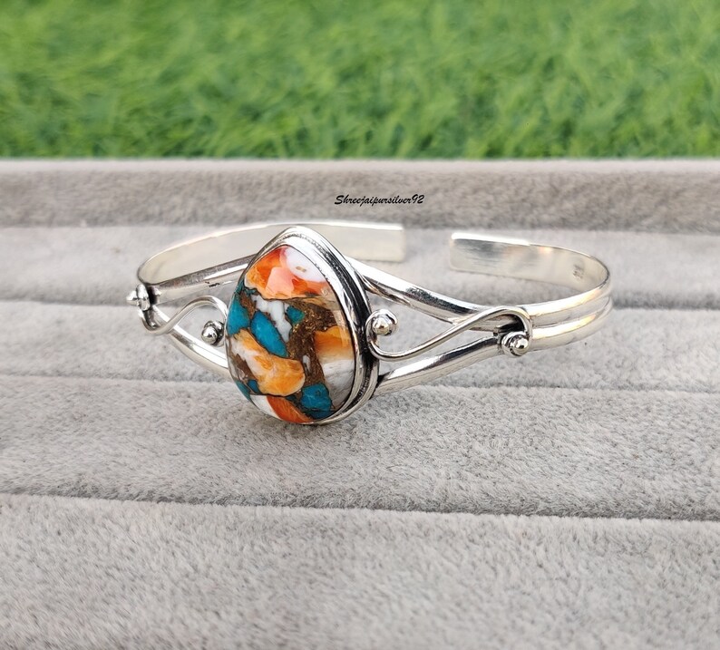 May include: A silver cuff bracelet with a teardrop-shaped turquoise stone set in the center. The stone is a mix of orange, blue, and white colors. The bracelet has a delicate, swirling design.