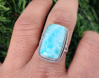 Könnte beinhalten: Ein silberner Ring mit einem rechteckigen blauen und weißen Edelstein. Der Ring wird an einem Finger getragen.