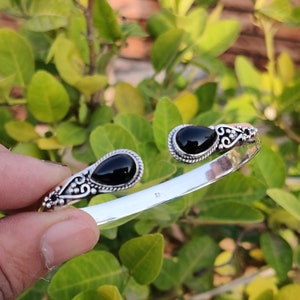 Puede incluir: Un brazalete de plata con dos piedras preciosas ovaladas negras. El brazalete tiene un diseño delicado y adornado.