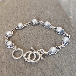 Puede incluir: Un brazalete de plata con cierre de palanca y detalles de perlas blancas.