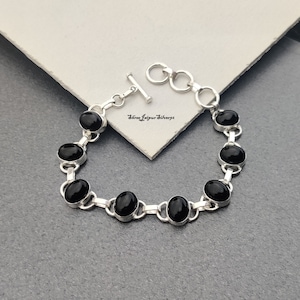 May include: A silver bracelet with oval black gemstones. The bracelet has a toggle clasp and is displayed on a grey surface. The gemstones are set in a silver link chain.